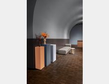 Minimalistisch gestalteter Innenraum mit abgerundeter Decke in Grau, einer dunkelbraunen Holzbodenfläche und schlichter Möblierung. Im Vordergrund befinden sich drei farbige Sockel mit einer Vase voller Trockenblumen, daneben stehen eine graue Polsterbank und ein kleiner Beistelltisch.