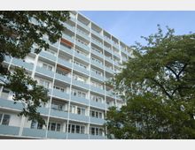 Mehrgeschossiges Wohngebäude mit einer Fassade aus hellblauen Balkonen und weißen Trägerkonstruktionen, umgeben von grünen Bäumen vor einem blauen Himmel.