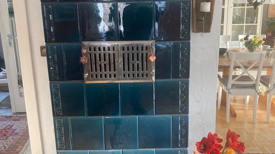 Ein dunkelblau glasierter Kachelofen mit rechteckigen Kachelfeldern und einem metallenen Lüftungsgitter in der Mitte, dekoriert mit kleinen Anhängern. Oben auf dem Ofen stehen Keramikkrüge, daneben befindet sich eine Wandkerze, und rechts davor ein Strauß bunter Blumen in einer Glasvase.
