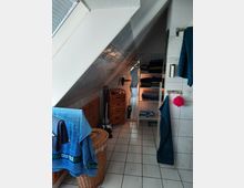 Badezimmer mit Dachschräge, gefliesten Wänden und weißem Fliesenboden. Im Raum befinden sich ein Wäschekorb aus Korbgeflecht, ein Handtuchhalter mit blauen Handtüchern, ein kleiner Schrank mit Schubladen und ein Regal mit gefalteten Handtüchern.