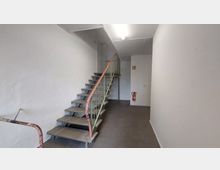 Ein einfacher Innenraum mit einer Metalltreppe auf der linken Seite, die zu einem oberen Stockwerk führt. Die Wände sind weiß gestrichen, der Boden ist grau, und an der gegenüberliegenden Wand hängt ein Feuerlöscher.