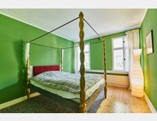 Schlafzimmer mit grün gestrichenen Wänden, einem Himmelbett mit geschnitzten Holzpfosten und kariertem Bettbezug. Der Raum hat Holzboden, ein großes Fenster mit weißen Vorhängen und eine stehende Papierlampe in der Ecke.