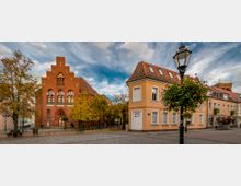 Städtischer Platz mit einem roten Backsteingebäude im Stil der Neogotik links im Bild und einem gelb gestrichenen Gebäude mit roten Dachziegeln rechts. Im Vordergrund stehen ein Baum mit gelbem Herbstlaub und eine Straßenlaterne mit Blumenschmuck.