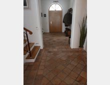 Flur eines Wohnhauses mit terrakottafarbenem Fliesenboden, einer Holztreppe links, einer Holztür mit halbrundem Glasfenster geradeaus und einigen Kleidungsstücken sowie Schuhen an der rechten Wand. Im Vordergrund steht eine Zimmerpflanze in einem Topf.