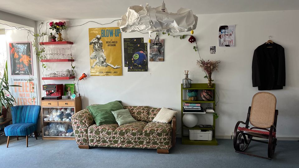 Eclectic living room with a patterned sofa, a blue upholstered chair, and a cane rocking chair on carpeted flooring. White walls are decorated with posters, hanging plants, and shelves holding glassware and small objects, with a homemade pendant lamp suspended from the ceiling.