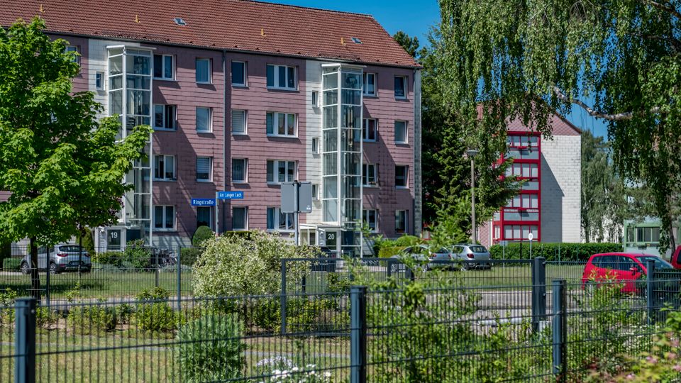 Mehrstöckiges Wohngebäude mit rosa Fassade und rotem Satteldach, mit verglasten Treppenhausaufgängen und mehreren Fenstern. Davor liegen eine Grünfläche mit Bäumen und Sträuchern sowie ein Zaun und geparkte Autos; im Hintergrund ist ein weiteres Gebäude mit roten Rahmen zu sehen.