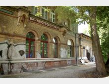 Backsteingebäude mit der Aufschrift „Bahnhofsgaststätte“, roten Fensterrahmen und Rundbogenfenstern, umgeben von Bäumen. Die Fassade ist teilweise mit Graffiti bedeckt, und der Gehweg davor besteht aus Kopfsteinpflaster.