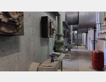 Industrieanlage mit Rohren, Ventilen und Maschinengehäusen in einem technischen Raum mit metallischen und betonierten Wänden. Im Vordergrund ein Steuerventil, im Hintergrund größere Rohrleitungen und Tanks.