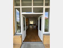 Blick durch geöffnete weiße Doppeltüren in einen langen Raum mit Holzboden, rechts und links befinden sich Wände mit Regalen und Möbeln; am Ende des Raumes sind Stühle und Fenster sichtbar. Der Eingangsbereich ist von gelbem Backstein eingefasst.