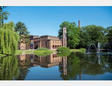 Backsteingebäude im Stil des Art déco mit hohen Fenstern und markantem Turm, gelegen an einem Teich mit Springbrunnen; umgeben von Bäumen und grünem Parkgelände bei klarem Himmel.
