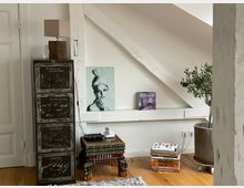 Wohnzimmerbereich in einem Dachgeschoss mit sichtbaren weißen Holzbalken. Zu sehen sind ein Metallaktenschrank, ein kleiner verzierter Holztisch mit Telefon, ein dekorativer Olivenbaum im Topf, ein Bücherstapel und gerahmte Kunstwerke an der Wand.