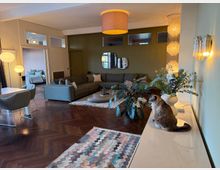 Modern open-plan living room with dark herringbone wood floors, an L-shaped gray sectional around a low oval coffee table, and pendant and wall lights. A white sideboard with vases and a large plant sits beside a geometric rug, with a doorway leading to a bedroom in the background.