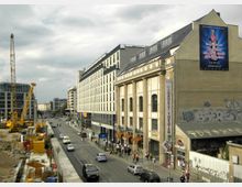 Straßenszene in einer Stadt mit moderner und historischer Architektur, rechts ein gelbes Gebäude mit hohen Fenstern und der Beschriftung „Kabarett Theater“; links eine Baustelle mit einem hohen Kran. Autos und Fußgänger beleben die Straße.