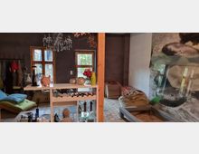 Ein gemütlicher, rustikaler Innenraum mit dunklen Wänden und Steinboden. Im Vordergrund steht ein kleiner Tisch mit Dekoration, darunter Vasen und Kerzenhalter, darüber hängt ein Kronleuchter, während im Hintergrund Sitzmöbel, Kissen und dekorative Elemente wie ein Blumenstrauß sichtbar sind.