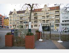 Straßenszene in einer deutschen Stadt mit einem mehrstöckigen, beige-gelben Wohngebäude im Gründerzeitstil im Hintergrund, das dekorative Fassaden und Balkone aufweist. Im Vordergrund sind ein kleiner Zaun, Blumenkästen aus rotem Backstein, Sonnenschirme sowie eine Bushaltestelle sichtbar.