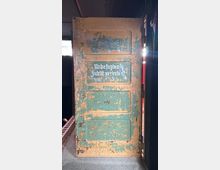 Alte, abgenutzte Holz- oder Metalltür mit abblätternder grün-beiger Farbe und mehreren eingelassenen Feldern, beschriftet mit „Unbefugten Zutritt verboten!“. Die Tür steht in einem dunklen Innenraum, seitlich fällt Licht durch eine Öffnung.