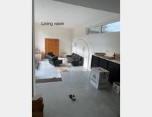 Wohnzimmer mit hellen Wänden, grauem Betonboden und moderner Einrichtung. Im Raum befinden sich schwarze Ledersessel, ein bogenförmiger Stehlampenschirm, eine Holzkommode und Umzugskartons; große Fenster lassen Tageslicht herein.