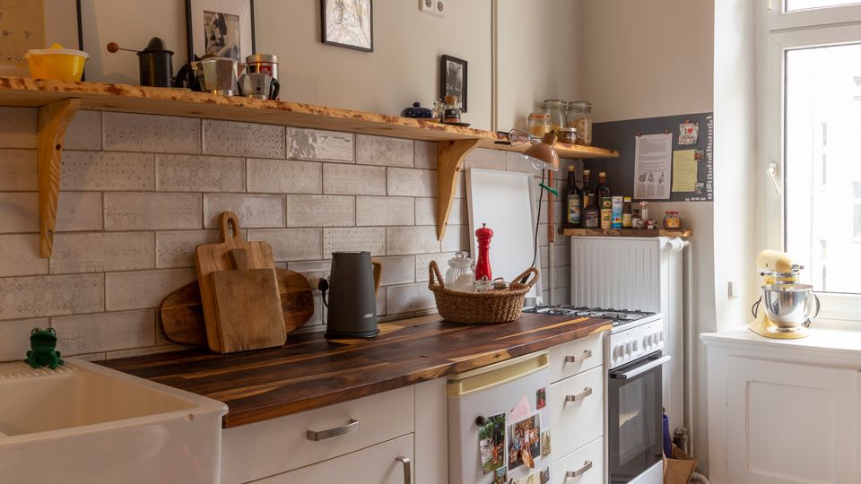 Gemütliche Küche mit weißer Unterschrankzeile und dunkler Holzarbeitsplatte, gefliestem Wandspiegel im Ziegelmuster und offenem Holzregal mit Gläsern und Küchenutensilien. Rechts sorgt ein hohes Fenster für Tageslicht; daneben stehen Herd und ein kleiner Kühlschrank, der Boden ist mit dekorativen Ornamentfliesen ausgelegt.