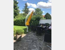 Garten mit einer gepflasterten Terrasse, auf der ein Esstisch mit schwarzen Rattan-Stühlen steht; im Hintergrund eine grüne Hecke, Bäume und ein gelber Sonnenschirm an einem Bogenständer.