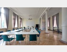 Ein großer, heller Seminarraum mit hohen Decken, verzierten Stuckelementen und weißen Säulen im klassizistischen Stil. Der Raum ist mit U-förmig angeordneten Tischen und blauen Stühlen ausgestattet, hat große Fenster mit bodenlangen Vorhängen und einen hölzernen Parkettboden.