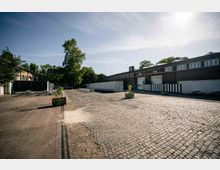 Ein großer, gepflasterter Hof mit vereinzelten Holzpflanzkübeln und Tischen auf Betonflächen, umgeben von Bäumen und niedrigen Backsteingebäuden mit Garagentoren und Fenstern. Im Hintergrund sind Container und eine sonnige, klare Himmelsszene zu sehen.