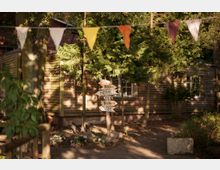 Holzhütte in einem bewaldeten Außenbereich mit einem Wegweiser aus Holz, der in verschiedene Richtungen zeigt, darunter „Toiletten“, „Paradies“ und „Biergarten“. Im Vordergrund hängen bunte Wimpel, und Bäume sowie Sträucher umgeben die Szene.