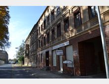 Straßenansicht eines historischen Gebäudes mit einer aufwendig verzierten Fassade und großen, dunklen Fenstern. Im Erdgeschoss befindet sich ein Geschäft mit der Beschilderung „Fleischerei Berg“, dessen Eingangsbereich mit braunen Fliesen verkleidet ist.