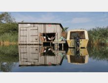 Holzgebäude auf Stelzen am Ufer eines ruhigen Gewässers, mit einem gelben Boot mit Außenbordmotor daneben; im Hintergrund sind Schilf und Bäume sichtbar. Auf der Terrasse des Gebäudes hängt eine Hängematte, in der eine Person liegt.
