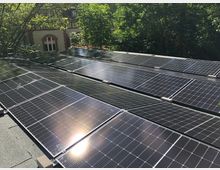 Solaranlage auf einem Flachdach, bestehend aus mehreren Reihen schwarzer Solarmodule. Im Hintergrund ist ein Gebäude mit weißen und roten Fassaden sowie umgebende grüne Bäume sichtbar.