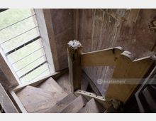 Holztreppe in einem alten Gebäude mit abgenutzten Stufen und einem beschädigten Holzgeländer. Rechts daneben eine verputzte Wand mit Wasserflecken, links ein Fenster mit Glasstäben, durch das Tageslicht einfällt.