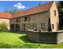 Ein zweistöckiges Gebäude aus Backstein im ländlichen Stil mit roten Ziegeldächern und Holzläden an den Fenstern. Im Vordergrund befindet sich eine Holzterrasse mit Blumenkübeln, umgeben von einer Rasenfläche und Bäumen im Hintergrund.
