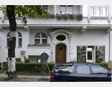Wohnhausfassade mit weißer Putzfassade im Stil der Gründerzeit, einem rundbogigen Holzeingang, dekorativen Fenstern und Balkon mit Blumenkasten. Vor dem Gebäude stehen ein Baum, ein parkendes Auto und ein Verkehrsschild.