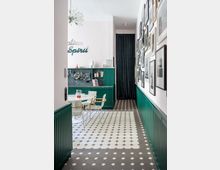 Schmaler Innenraum mit gemusterten Fliesen in Weiß und Grau, grünen Wandverkleidungen und einer ausgestellten Fotogalerie auf der rechten Seite. Links befindet sich ein kleiner Essbereich mit einem runden Tisch und Stühlen, sowie eine Wandküche mit offenen Regalen und Kochutensilien.