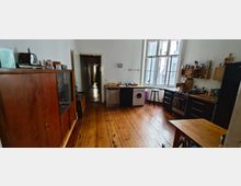 Wohnküche in einer Altbauwohnung mit Holzfußboden und hohen Decken. Links steht ein antikes Holzsideboard, rechts befinden sich Küchengeräte wie ein Herd, eine Spüle und eine Waschmaschine, vor einem großen Fenster mit Blick in einen Innenhof.