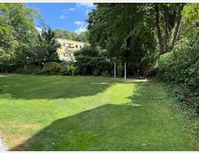 Grüne Rasenfläche in einem Garten, umgeben von dichten Büschen und hohen Bäumen. Im Hintergrund ist ein gelbes Wohngebäude mit mehreren Fenstern und Balkonen zu sehen.