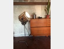 Innenraum mit rauer Betonwand und einem hölzernen Sideboard, auf dem kleine Pflanzen und Kameraobjektive stehen. Daneben steht ein großer, industrieller Scheinwerfer auf einem Stativ mit sichtbarem Kabel.