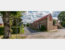 Straßenszene in einem Dorf mit einer gepflasterten und einer asphaltierten Straße, flankiert von Häusern aus Ziegelmauerwerk, einem großen Baum und gepflegten Hecken. Im Hintergrund sind weitere Gebäude und ein bewölkter Himmel sichtbar.