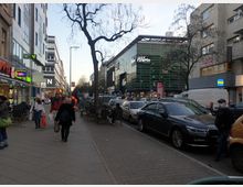 Straßenszene mit Fußgängerzone auf der linken Seite und parkenden Autos rechts entlang der Fahrbahn. Im Hintergrund ein modernes Gebäude mit Glasfassade und der Aufschrift „Wilma“, flankiert von Geschäften, Cafés und weiteren Gebäuden.