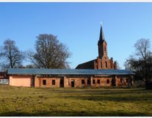 Backsteingebäude mit flachem Blechdach, davor eine Wiese, im Hintergrund eine rote Kirche mit einem hohen Spitzturm und umliegenden Bäumen unter klarem blauen Himmel.