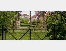 Ein Garten mit grünem Rasen, umgeben von Bäumen und Sträuchern, im Hintergrund Wohnhäuser mit roten Ziegeldächern und Fenstern; im Vordergrund ein Metallzaun.