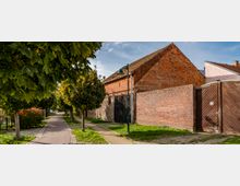 Eine ruhige Straße mit gepflastertem Gehweg, gesäumt von Bäumen und umgeben von Ziegelbauwerken, darunter eine alte, teilweise verwitterte Backsteinmauer und ein Gebäude mit ziegelrotem Giebel. Im Vordergrund steht eine grüne Straßenlaterne.