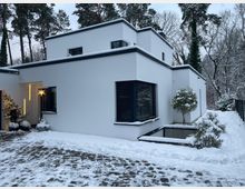 Modernes, weiß verputztes Flachdachhaus mit kubischer, gestufter Architektur und großen Fenstern mit dunklen Außenjalousien. Im verschneiten Vorgarten liegen ein gepflasterter Weg, niedrige Mauern und Sträucher; dahinter steht ein Wald aus hohen Bäumen.