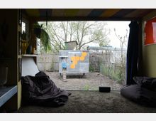 Ausblick aus einem Innenraum mit Sitzkissen auf einen Garten mit einem Hühnerverschlag. Der Verschlag ist aus Holz und Metall gebaut, mit einer Leiter und einer Verzierung aus gelben Wabenformen; im Hintergrund sind Bäume und ein Holzzaun zu sehen.