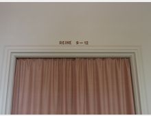 Türöffnung mit einem beigefarbenen Vorhang, der vollständig geschlossen ist. Über der Tür steht in einfacher Schrift „REIHE 9–12“.