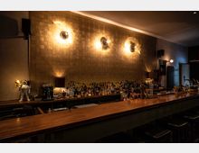 Bar mit einer langen Theke aus Holz, dahinter eine gold schimmernde Wand mit drei runden Wandlampen, die ein warmes Licht abstrahlen. Im Hintergrund stehen zahlreiche Flaschen Spirituosen, Gläser und Barutensilien, links ist eine kleine Astronautenfigur als Dekoration zu sehen.