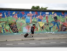 Ein Abschnitt der Berliner Mauer mit bunten Graffiti und Zeichnungen, darunter der Schriftzug „Cool for the East“. Auf dem Gehweg davor geht eine Person mit einem Rollkoffer entlang.