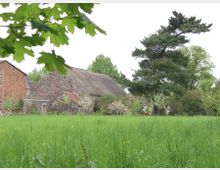 Eine ländliche Szene mit einer aus Natursteinen errichteten Scheune und einem roten Backsteingebäude im Hintergrund, umgeben von blühenden Sträuchern, einem großen Nadelbaum und einer grünen Wiese im Vordergrund. Blattwerk eines Baumes hängt teilweise ins Bild.