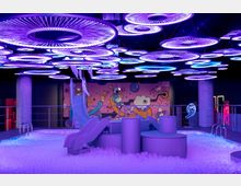 Ein Indoor-Spielplatz mit futuristischer Beleuchtung in lila und blau. Die Szenerie umfasst eine Kinderrutsche, eine Ballgrube und eine Wand mit bunten Cartoon-Grafiken im Hintergrund.