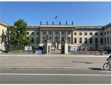 Eine repräsentative, klassizistische Fassade der Humboldt-Universität zu Berlin, mit Säulen, Statuen und einer zentralen Statue im Eingangsbereich. Vor dem Gebäude befinden sich ein gepflasterter Platz, einige Werbebanner und eine Straße mit vorbeifahrenden Personen.