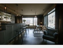 Modernes Innenraumdesign mit einer dunklen Bar auf der linken Seite, hohen Hockern, und einer Sitzecke mit einem großen Fenster, das Tageslicht und den Blick auf eine städtische Umgebung bietet. Der Raum ist mit dunklen Holzböden, hängenden Lampen und Möbeln in neutralen Farben ausgestattet.